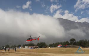 Everest Helicopter Tour -Red and white helicopters parked or hovering on a wide grassy field with dramatic low-hanging clouds rolling over a massive mountain peak in the background.